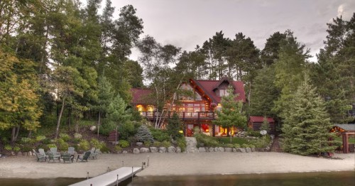 A lakeside cabin surrounded by trees, featuring a sandy beach and a wooden dock. Warm lights glow from the windows.
