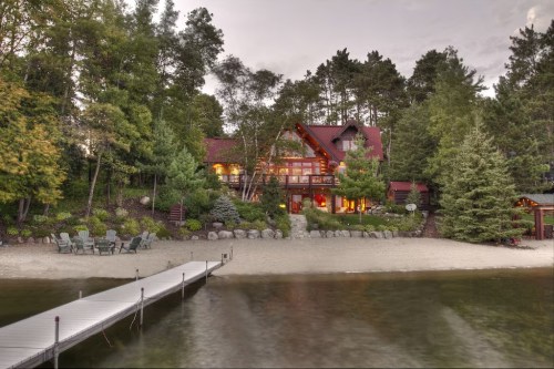 A lakeside cabin surrounded by trees, with a sandy beach and a wooden dock leading to the water.