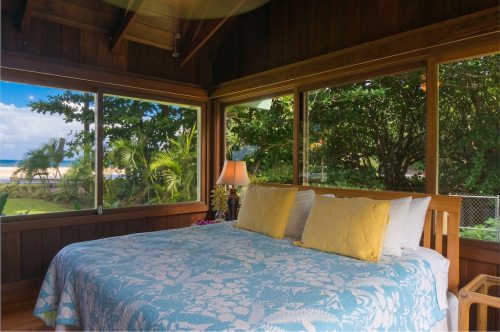 Cozy bedroom with a blue and white quilt, large windows showcasing greenery and a beach view.