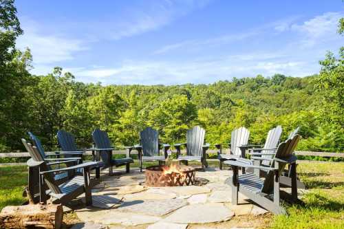 A circular stone fire pit surrounded by wooden chairs, set against a backdrop of lush green trees and a clear blue sky.