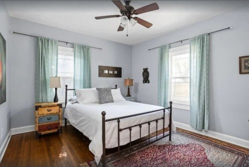A cozy bedroom with a metal bed, light blue walls, curtains, a colorful nightstand, and a patterned rug.