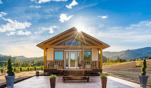 A modern cabin with a large front porch, surrounded by mountains and vineyards under a bright blue sky.