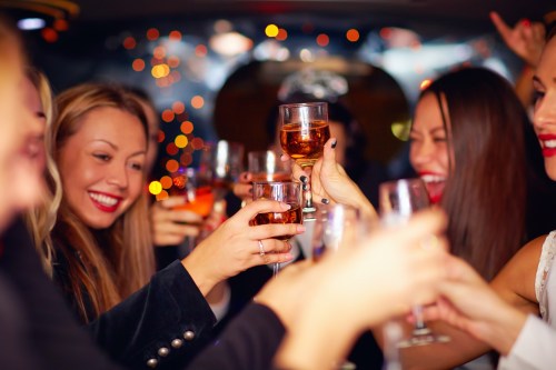 A group of smiling friends toasting with drinks in a festive setting, surrounded by colorful lights.