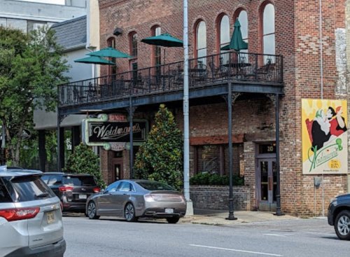 A brick building with a balcony, green umbrellas, and a colorful mural on the side, viewed from the street.