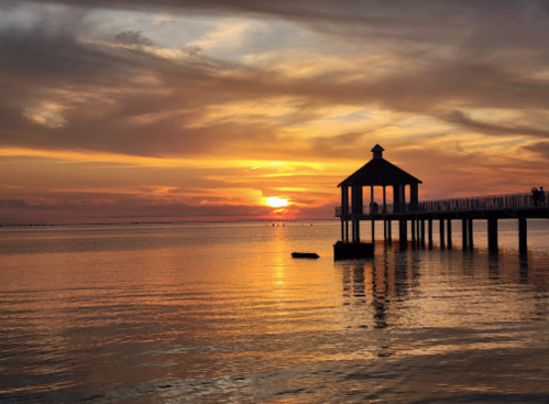 A serene sunset over calm waters, with a pier and a small gazebo silhouetted against the colorful sky.