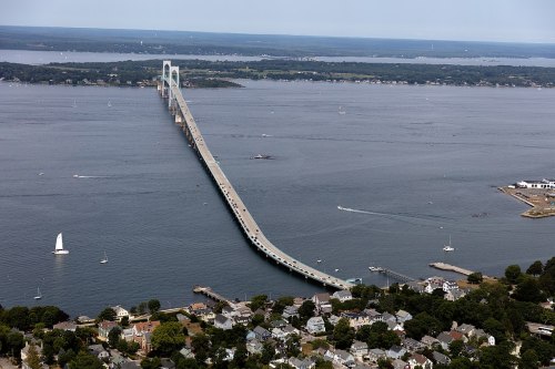 Aerial view of a long bridge spanning a wide body of water, with boats and a coastal town visible below.