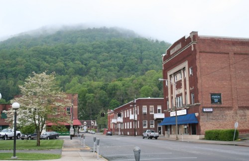 A small town street with brick buildings, a flowering tree, and a misty green mountain in the background.
