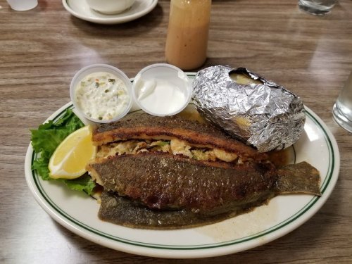 A plate with a whole fried fish, lemon, a baked potato wrapped in foil, and two dipping sauces on the side.