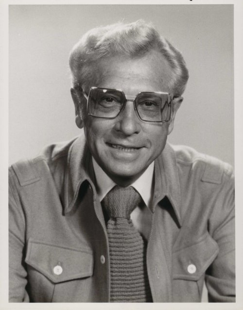 Black and white portrait of a smiling man with glasses, wearing a light jacket and tie, against a plain background.