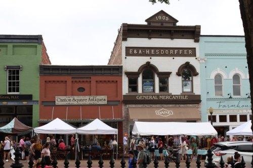 Historic storefronts line a street, featuring shops and tents with people browsing at an outdoor market.