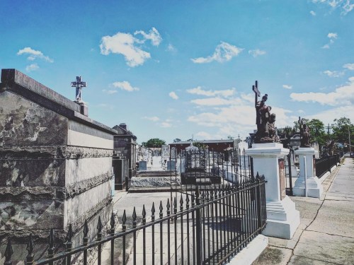 A cemetery scene featuring ornate tombstones, a statue, and a clear blue sky with scattered clouds.