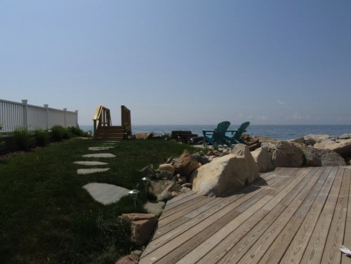 A serene waterfront scene featuring a grassy area, wooden deck, rocks, and blue chairs overlooking the water.