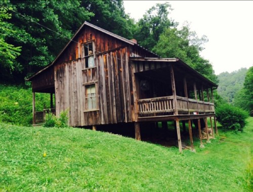 A rustic wooden house on stilts, surrounded by lush green grass and trees in a serene, hilly landscape.