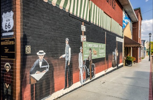 Mural depicting historical scenes along Route 66, featuring people and vintage signage on a brick wall.