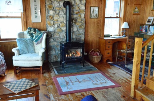 Cozy interior with a stone fireplace, armchair, wooden furniture, and a rug, creating a warm, inviting atmosphere.