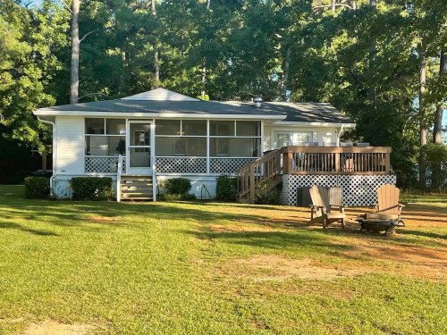 A cozy white house with a porch, surrounded by trees and a grassy area, featuring a fire pit and seating.