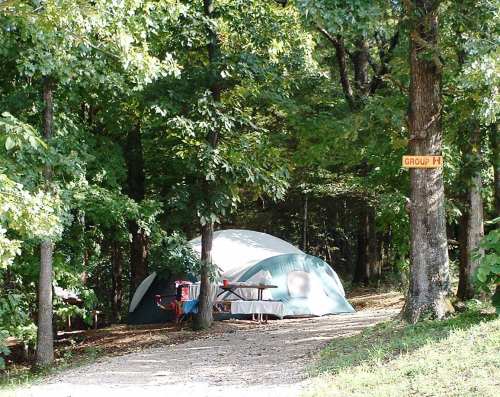A green tent set up in a wooded area, with a picnic table nearby and a sign labeled "Group H" on a tree.