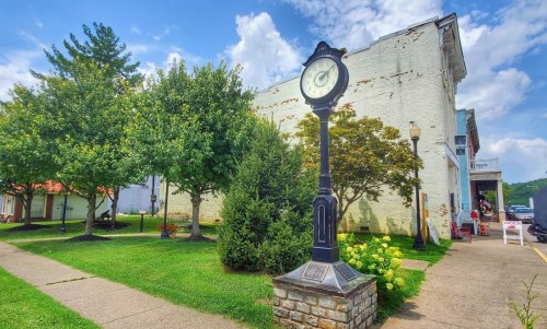 A vintage clock stands in a green park with trees and a brick building in the background under a blue sky.