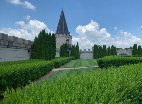 A castle-like structure with a tall spire surrounded by neatly trimmed hedges and a blue sky with fluffy clouds.