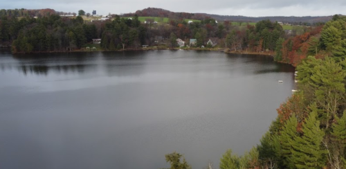 A serene lake surrounded by trees, with houses visible along the shore and autumn foliage in the background.