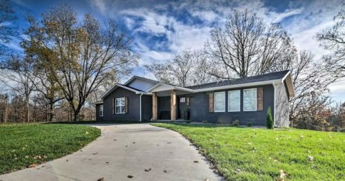 A modern single-story house with a curved driveway, surrounded by trees and a grassy lawn under a blue sky.