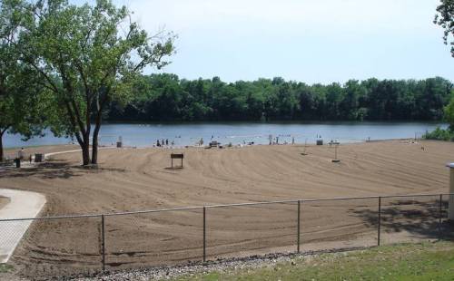 A sandy beach by a calm river, surrounded by trees, with people enjoying the water in the distance.