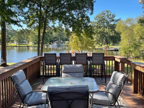 A lakeside deck with seating and a table, surrounded by trees and a view of the water and boats.