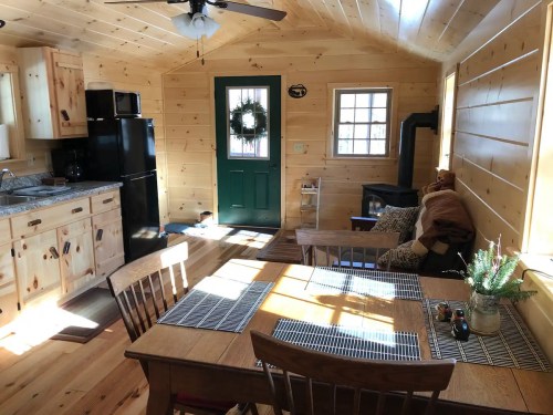 Cozy wooden cabin interior with a kitchen, dining table, and a green door, featuring natural light and rustic decor.