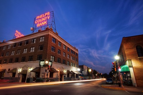A city street at dusk featuring the Phelps County Bank building with neon signage and light trails from passing cars.