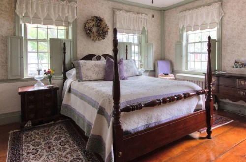 Cozy bedroom with a wooden bed, floral bedding, and light green walls, featuring windows and a decorative rug.