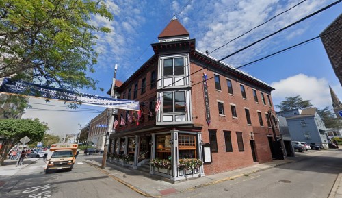 A brick building with a peaked roof, featuring flags and flower boxes, located on a street with clear skies.