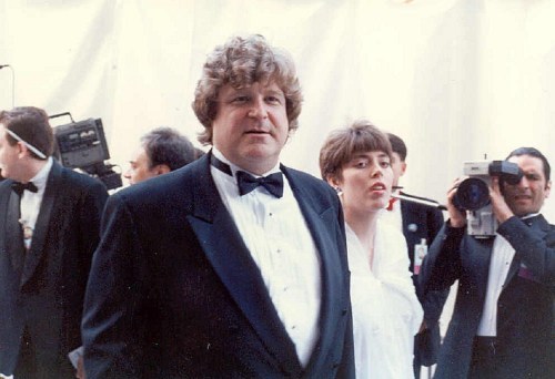 A man in a tuxedo stands with a woman in a white dress, surrounded by people at a formal event.