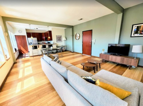 Bright, modern living room with a sofa, coffee table, and kitchen in the background, featuring large windows and wooden floors.