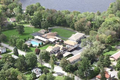 Aerial view of a resort with a pool, surrounded by trees and a river, featuring multiple buildings and green spaces.