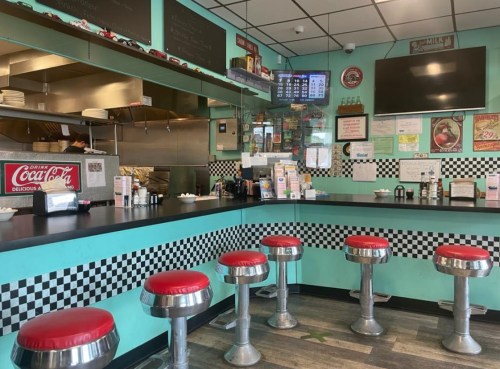 A retro diner interior with red stools, a checkered counter, and a menu board in a turquoise setting.