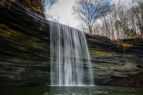 A serene waterfall cascading over layered rock formations into a calm pool, surrounded by trees and a cloudy sky.