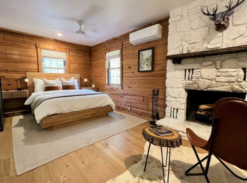 Cozy bedroom with wooden walls, a bed with white linens, a stone fireplace, and a rustic chair and table.