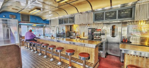 A retro diner interior with a long counter, red stools, and a man sitting alone, surrounded by vintage decor.