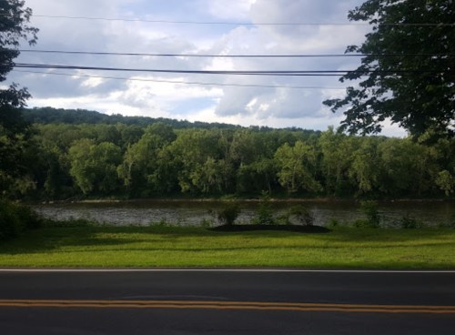 A serene river scene with lush green trees lining the bank, under a partly cloudy sky. A road runs in the foreground.