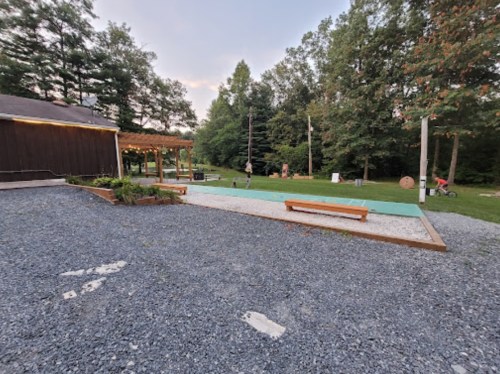 A gravel area with a green court, benches, and trees in the background, near a wooden building and a pergola.