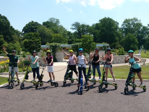 A group of eight people on scooters, smiling and waving in a sunny park setting.