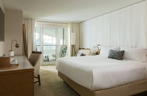 A modern hotel room featuring a large bed, desk, and balcony with a view, decorated in neutral tones.