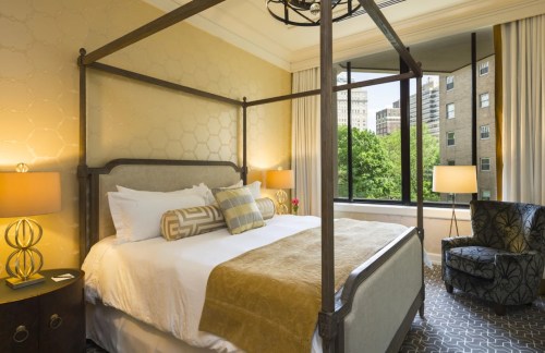 A stylish hotel room featuring a four-poster bed, cozy armchair, and large window with city views.