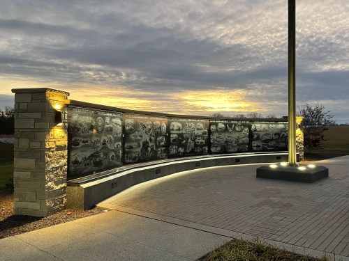 A memorial wall with engraved images, illuminated by lights, against a sunset sky.