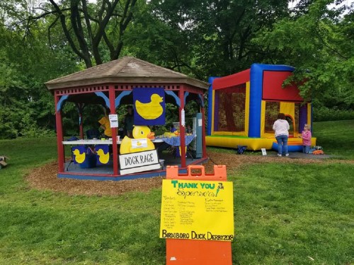 A colorful gazebo with a duck race sign and a bounce house nearby, set in a green park.
