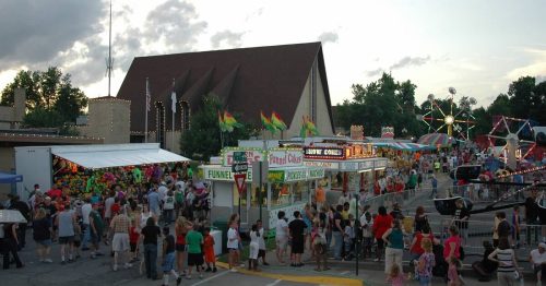 A lively fair scene with crowds, food stalls, games, and colorful rides under a cloudy sky.