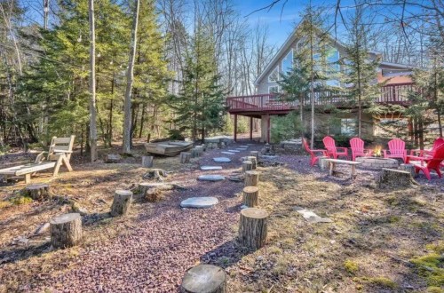 A serene outdoor space with a stone path, wooden seating, and red chairs surrounded by trees and a house in the background.