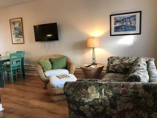 Cozy living room with floral couch, wicker chair, lamp, and TV, featuring a dining area with a blue table and chairs.