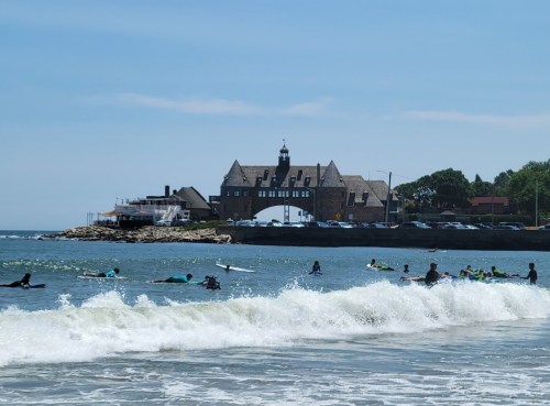 A beach scene with surfers in the water and a historic building in the background under a clear blue sky.
