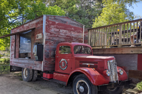 A vintage red truck converted into a rustic food stand, parked beside a wooden deck in a green outdoor setting.